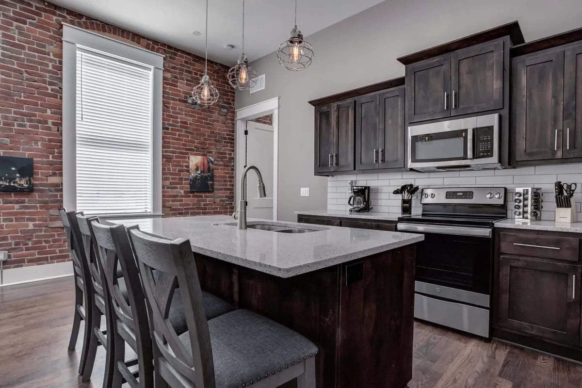 A spacious kitchen featuring a gleaming granite countertop, exposed original brick wall, and stylish industrial light fixtures. Stainless steel appliances line the space, and the inviting atmosphere is perfect for both cooking and entertaining. The wood floors add warmth and character, tying in the historic elements with modern touches.