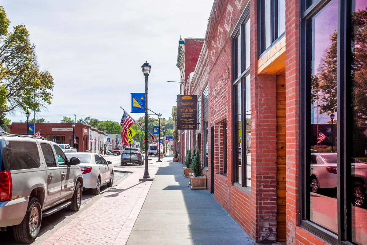 A picturesque street view of brick buildings, decorative street lamps, parked cars, and flags, highlighting bustling storefronts and the community atmosphere. The image evokes a sense of strolling, shopping, and enjoying neighborhood life.