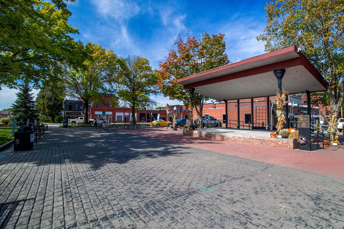 An open stage set against colorful trees and historic brick buildings, suggesting spaces for community gatherings, concerts, and seasonal events. The area feels lively yet accessible, perfect for families.
