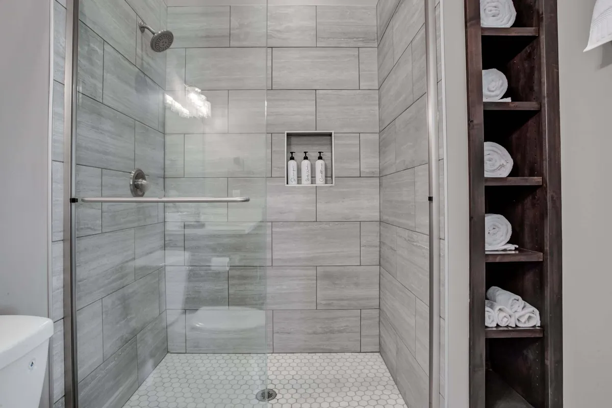 A spa-like bathroom featuring a glass shower enclosure, wall-mounted shelf with toiletries, neatly arranged towels, and sleek hexagonal floor tiles. The serene atmosphere is perfect for unwinding after a busy day.