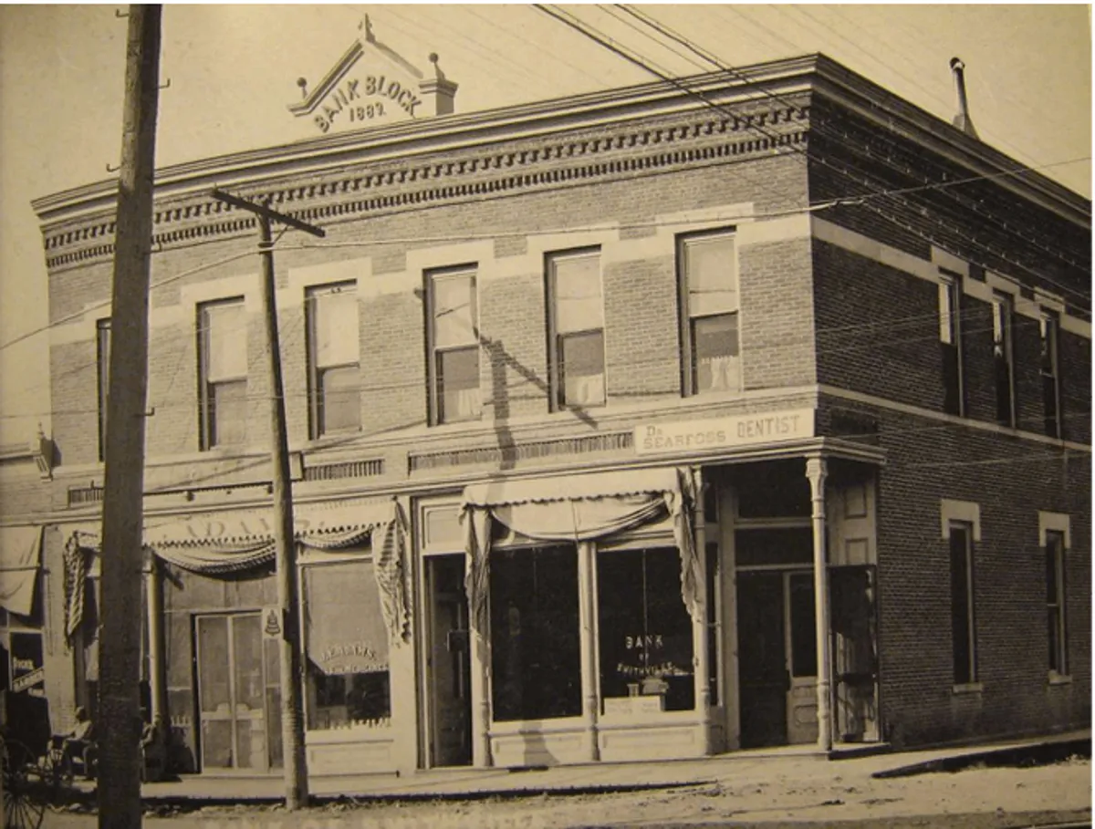 This image shows the historic Bank Block building with its distinctive brickwork, decorative awnings, and classic business signage. A dentist office sign is visible, and the scene captures the bustling activity of town residents socializing and going about daily life in the town square.