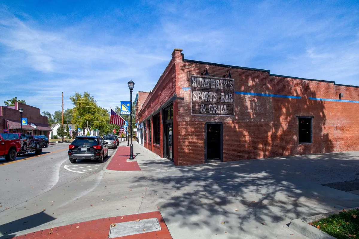 Features Humphrey's Sports Bar & Grill sign on a traditional brick façade overlooking a lively street scene with parked cars. The mood is social and energetic, hinting at communal events, casual dining, and nightlife.