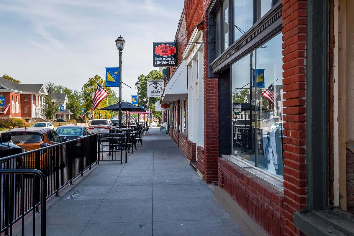 A lively downtown streetscape featuring charming brick storefronts, inviting outdoor cafe seating, and street lamps adorned with flags. The scene is bustling with diners enjoying al fresco meals and people strolling by, capturing the heart of Smithville's vibrant culinary scene.
