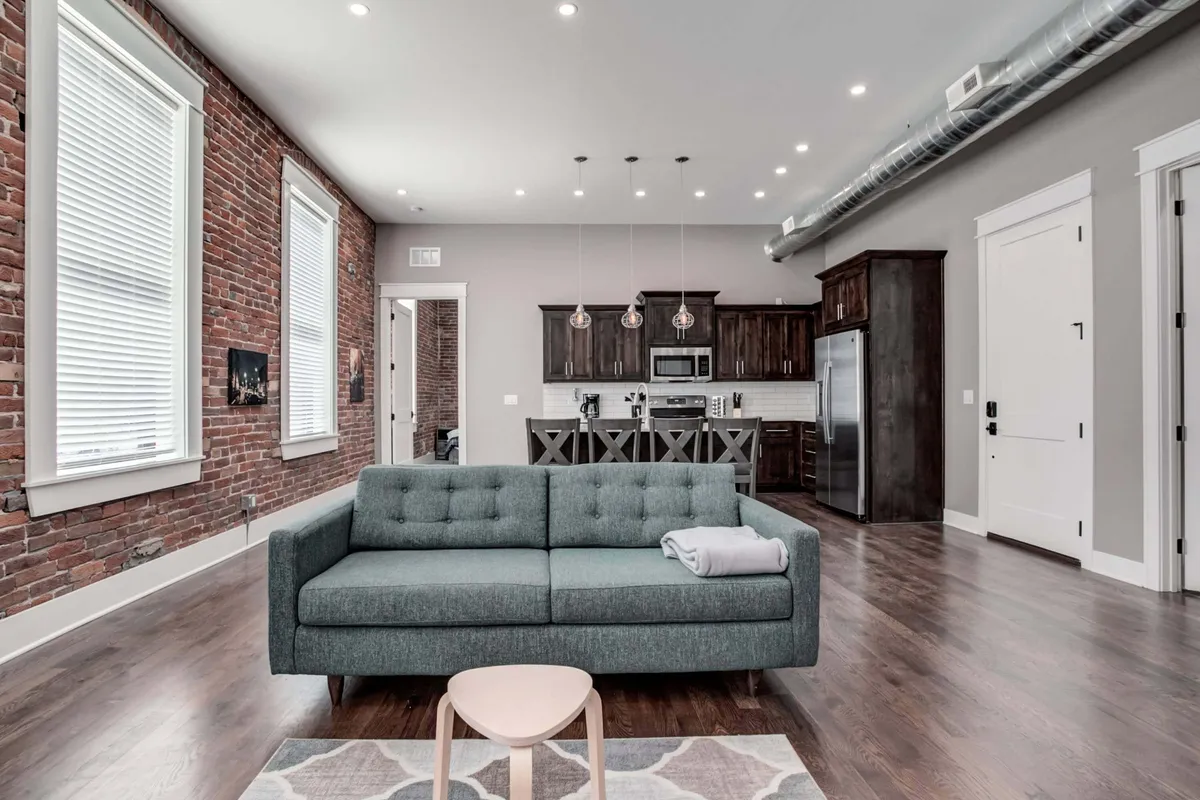 An open-plan living and kitchen area with a comfortable sofa, exposed brick wall, and sleek light fixtures. The kitchen is visible in the background, seamlessly integrated with the living space. It captures a sense of socializing, relaxation, and the blending of historic and modern elements.