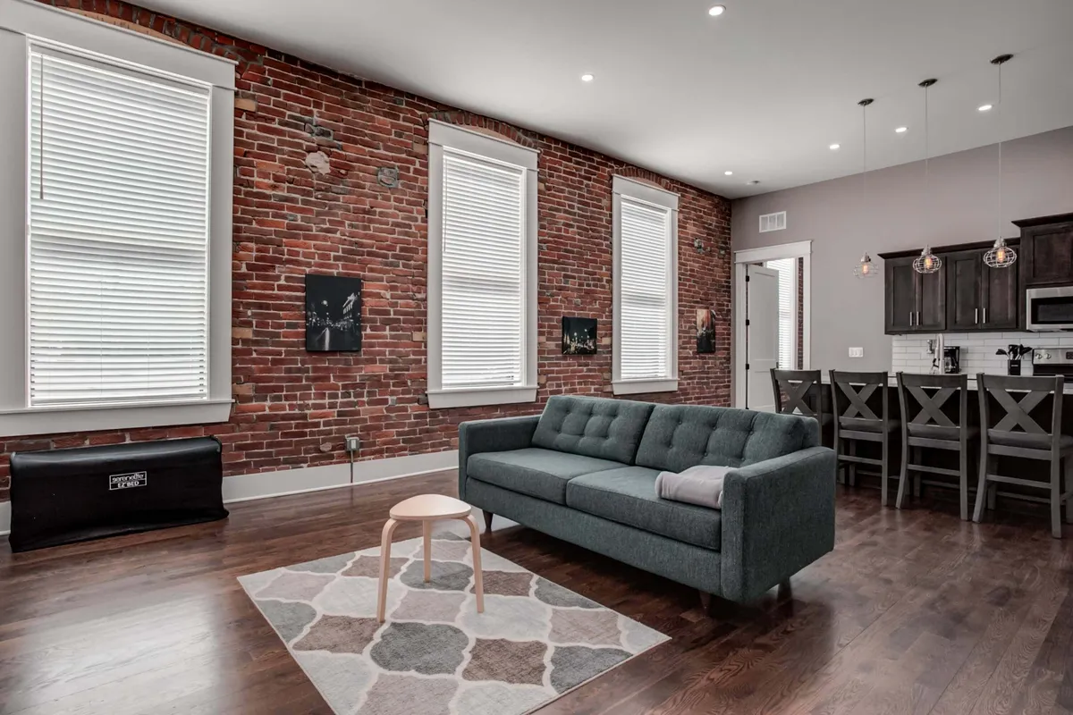 The living area is captured from a slightly different angle, highlighting a modern sofa, an exposed brick wall, kitchen space, and curated art pieces. The decor is high-end and visually cohesive, suggesting a space ideal for both relaxation and entertaining.
