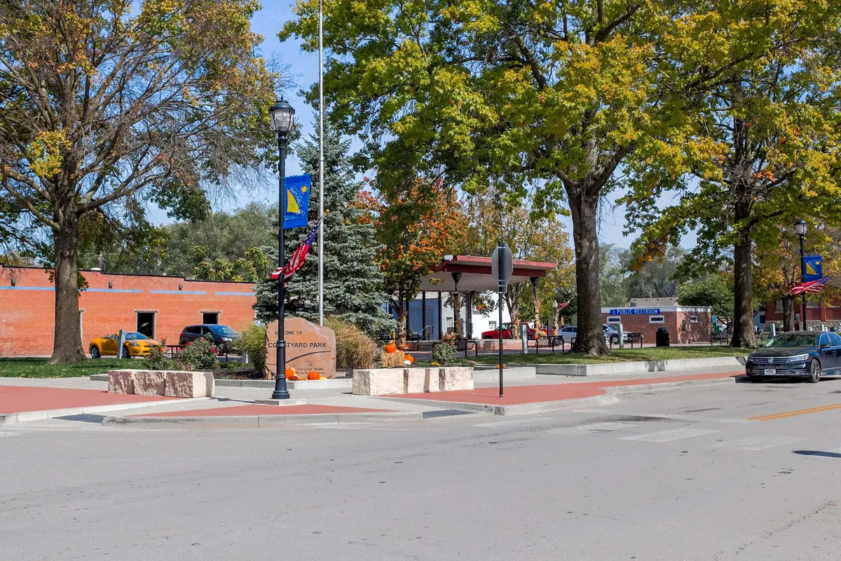 A picturesque view of Courtyard Park, with autumn-colored trees, lampposts, welcoming park benches, and a prominent park sign. The park is dotted with people walking and relaxing, creating a cheerful, inviting setting for outdoor gatherings or picnics.