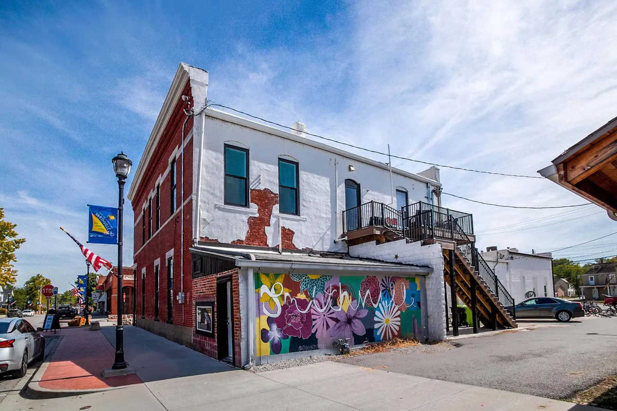 A dynamic exterior scene featuring an eye-catching mural, exterior stairway, lamp post, and colorful flags. Locals and visitors explore the area, taking in local art and participating in the energetic street life that characterizes Smithville’s creative side.