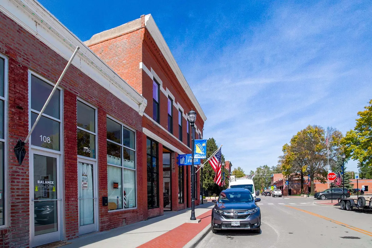 A lively street scene featuring a charming brick building facade, large storefront windows, a classic street lamp, and American flags fluttering along the sidewalk. The image captures pedestrians strolling past bustling local shops and enjoying the inviting outdoor atmosphere.