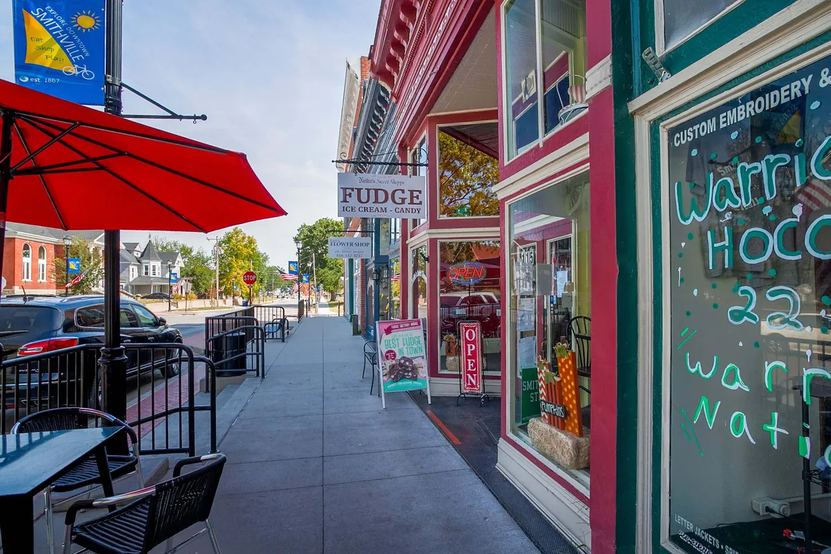 A colorful downtown streetscape with cheerful storefronts, a red umbrella shading outdoor seating, and a fudge shop's welcoming 'open' sign. People enjoy ice cream, shop for unique local goods, and relax under bright skies, giving the scene a festive, community-oriented feel.