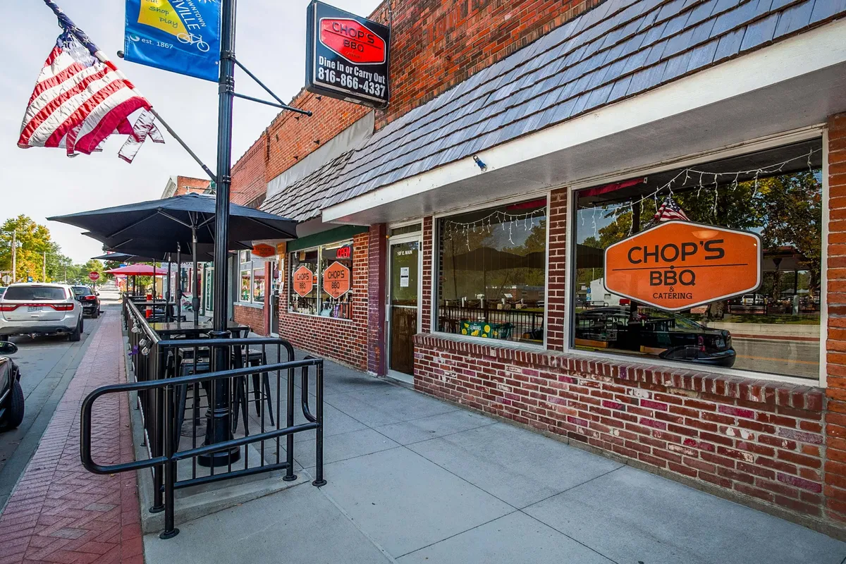 A welcoming restaurant facade with a prominent 'Chop's BBQ' sign, American flags, and plentiful outdoor seating. Diners are seen enjoying meals and conversations in a friendly, sociable setting, reflecting the town’s community spirit and culinary delights.