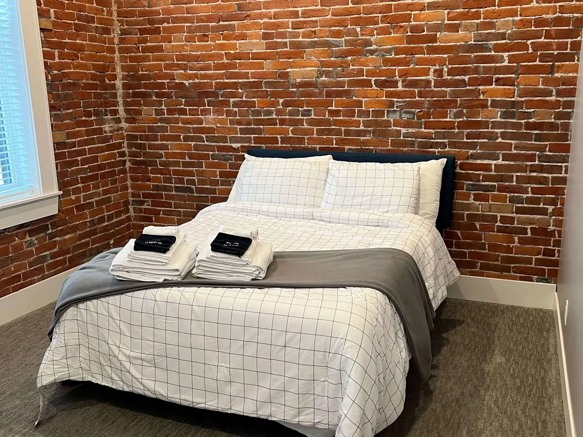 A stylish bedroom featuring an exposed brick wall, patterned bedding, and neatly arranged towels. Natural light spills into the room, adding to the cozy and modern feel, while the tasteful décor emphasizes both comfort and visual appeal.