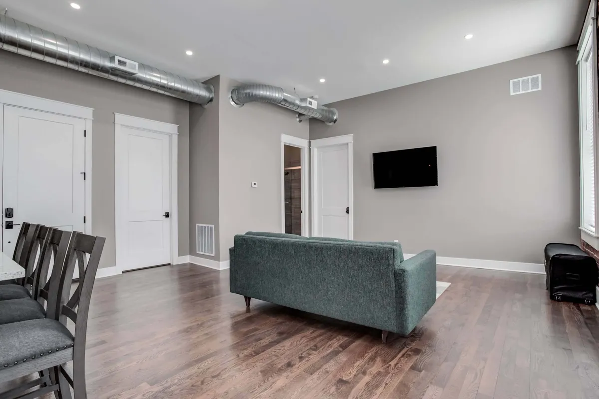 A calm, modern dining area adjacent to a green sofa, with a wall-mounted television and a stylish table setup. The space is designed for both intimate dinners and lively gatherings, and the wood flooring and inviting layout enhance the sense of comfort and style.