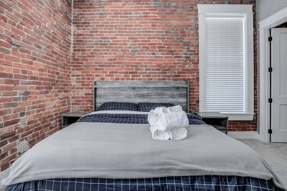 A tranquil bedroom with an exposed brick wall creates a warm backdrop for a wooden bed dressed in sumptuous linens. Folded towels are thoughtfully placed at the foot of the bed, and sunlight filters softly through a window, enhancing the inviting atmosphere.