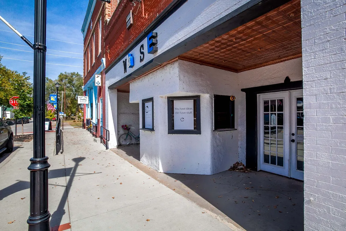 Depicts an inviting streetscape with business signage, entrance doors, a bicycle, and decorative planters. The setting suggests a space for casual dining, shopping, and community gatherings, with hints at local art and culinary experiences.