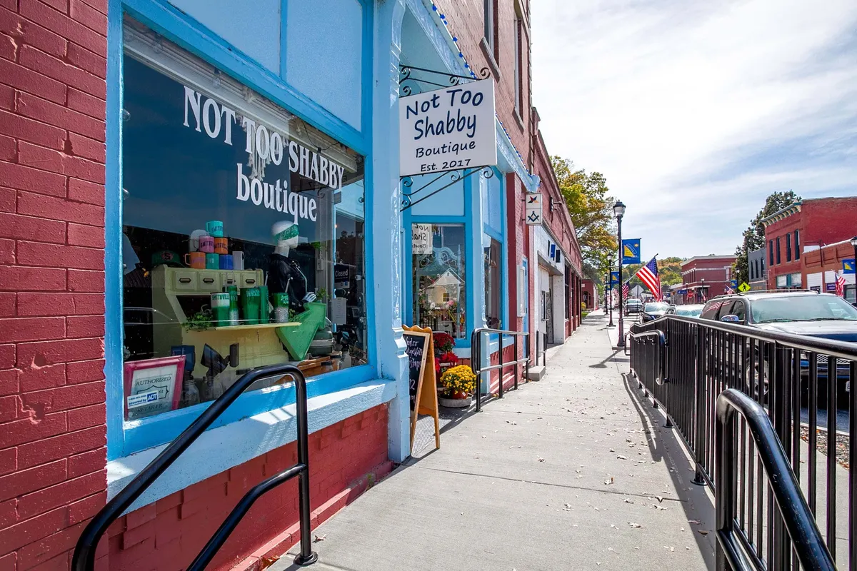 The image showcases a charming storefront sign with a detailed window display and flower pots lining the exterior. The scene is inviting, capturing the small-town vibe and the appeal of exploring local shops after a day out.